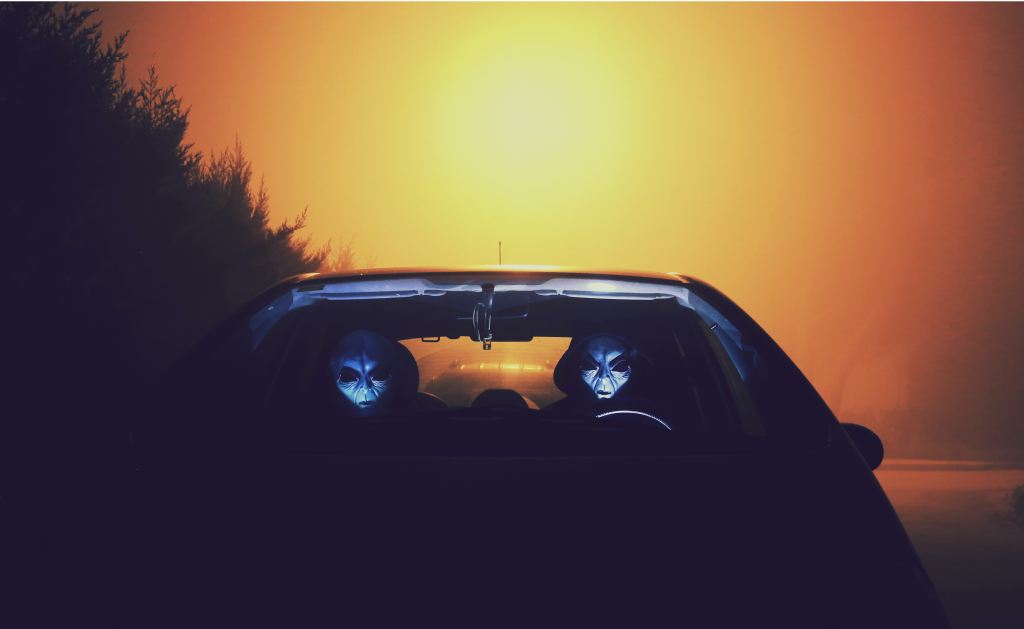 Two aliens in a car at night silhouetted against a hazy orange light 