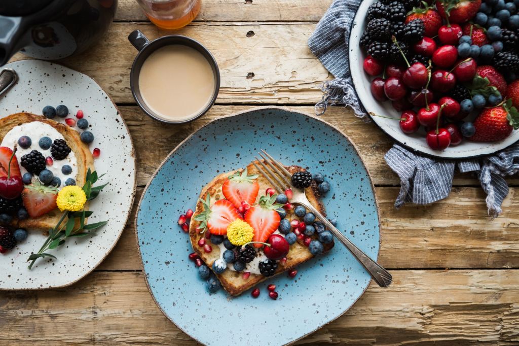 Overhead photo of toast and fruit on plates with a cup of tea and a bowl of berries and cherries on a rustic wooden table