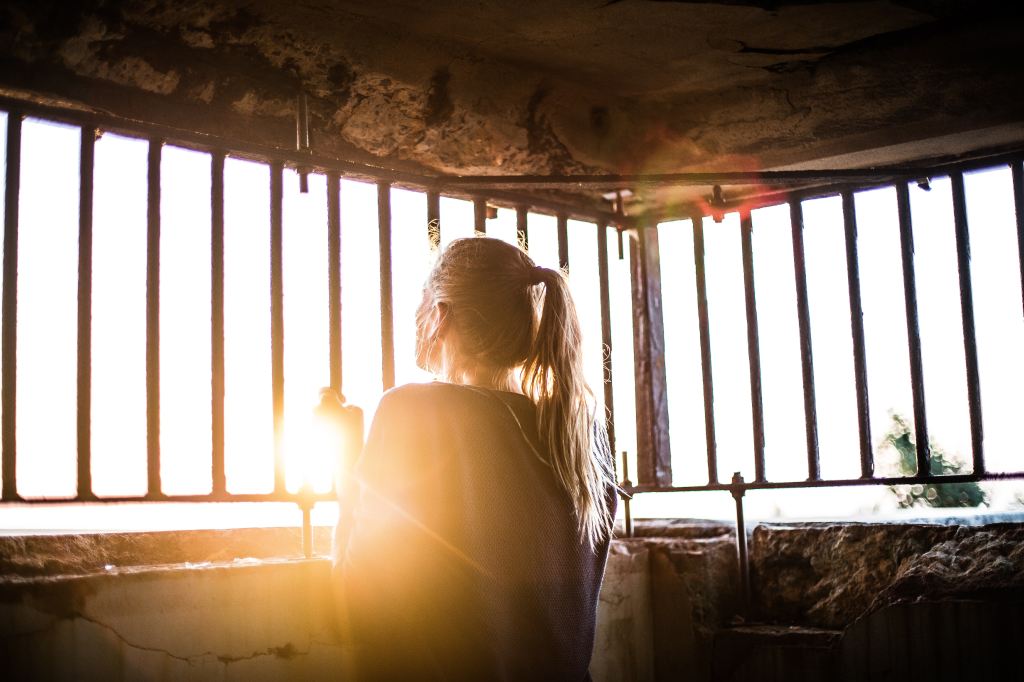 Woman with a long blonde ponytail looking out through bars toward the sun low on the horizon