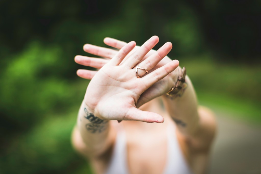 A woman is holding up her hands so that her face can't be seen. She's wearing a ring, a watch and there are tattoos on her wrists. The rest of the image is blurred.